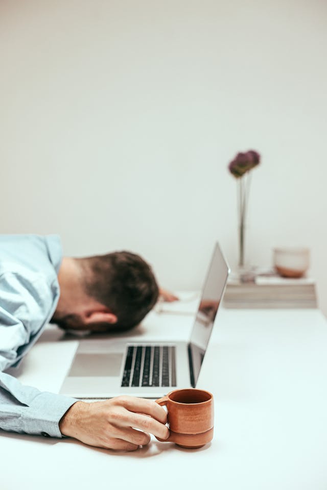 Tired man with head down, holding a coffee cup in front of an open laptop, symbolizing exhaustion and fleeting motivation.